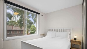 Un dormitorio con un ventanal grande, una cama blanca, una mesita de noche con lámpara y una pequeña mesita auxiliar de madera.
