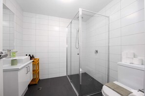 A modern bathroom with white tiles, a glass shower enclosure, a toilet, and a small wooden shelf with toiletries. - Two Bedroom Retreat Opposite Victoria Park (Abbotsford)