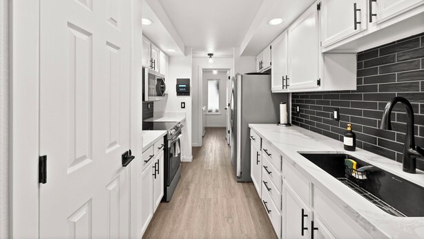 A narrow kitchen with white cabinetry, a black backsplash, and a stainless steel sink.