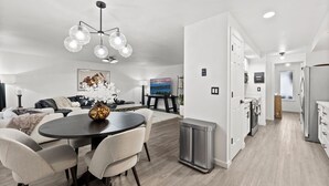 A modern kitchen with stainless steel appliances, a dining area with a round table and chairs, and a living room with a sofa and a painting on the wall.