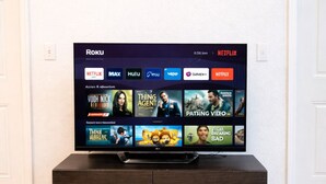 Een televisie op een donker houten voetstuk, tegen een witte muur, toont een Roku-interface met verschillende streamingdiensten en filmminiatuurafbeeldingen.