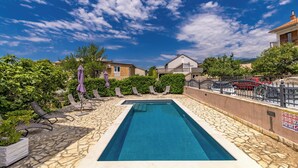 Ein rechteckiger Pool mit Steinplatten, umgeben von Liegestühlen und Sonnenschirmen, mit blauem Himmel und Wohnhäusern im Hintergrund.