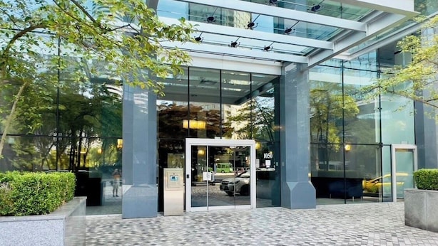 Ingresso moderno di un edificio con porte in vetro, pavimento piastrellato e vegetazione circostante.