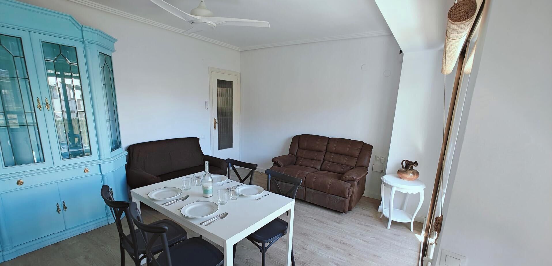 Un comedor con una mesa preparada para cuatro personas, un sofá marrón, un mueble azul y una mesita blanca con un adorno.