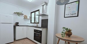A compact kitchen with white cabinets, a built-in oven, a stove, and a small round table with a plant. - Guest Ready - Beachside Flat in Puerto de Sagunto (El Port de Sagunt)