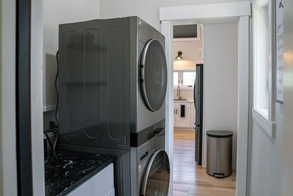 A compact laundry area with a front-loading washer and dryer, a stainless steel trash bin, and a kitchen visible in the background.