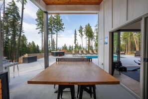 An outdoor dining area with a wooden table and chairs, a hot tub, and a poolside seating area with lounge chairs. - The Overlook at Flathead Lake - Wedding Venue (Lakeside)