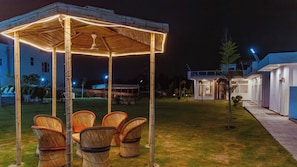 A gazebo with wicker chairs and a ceiling fan, surrounded by a grassy area and a building with a white exterior.