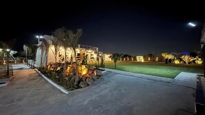 Un espacio exterior bien iluminado con piscina, palmeras y un edificio con ventanas iluminadas.