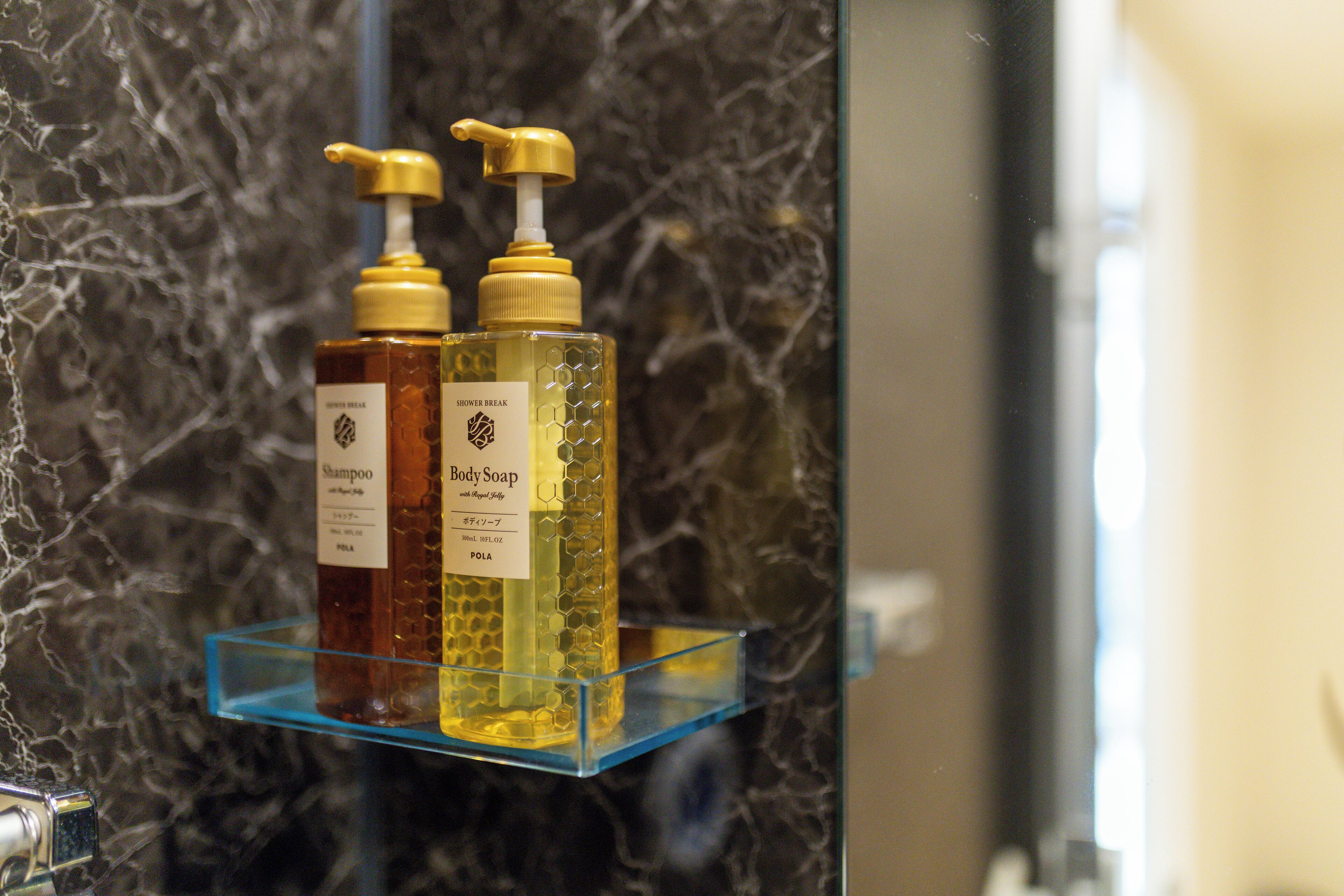 A bathroom with a marble wall and a glass shelf holding two bottles of shampoo and body soap.