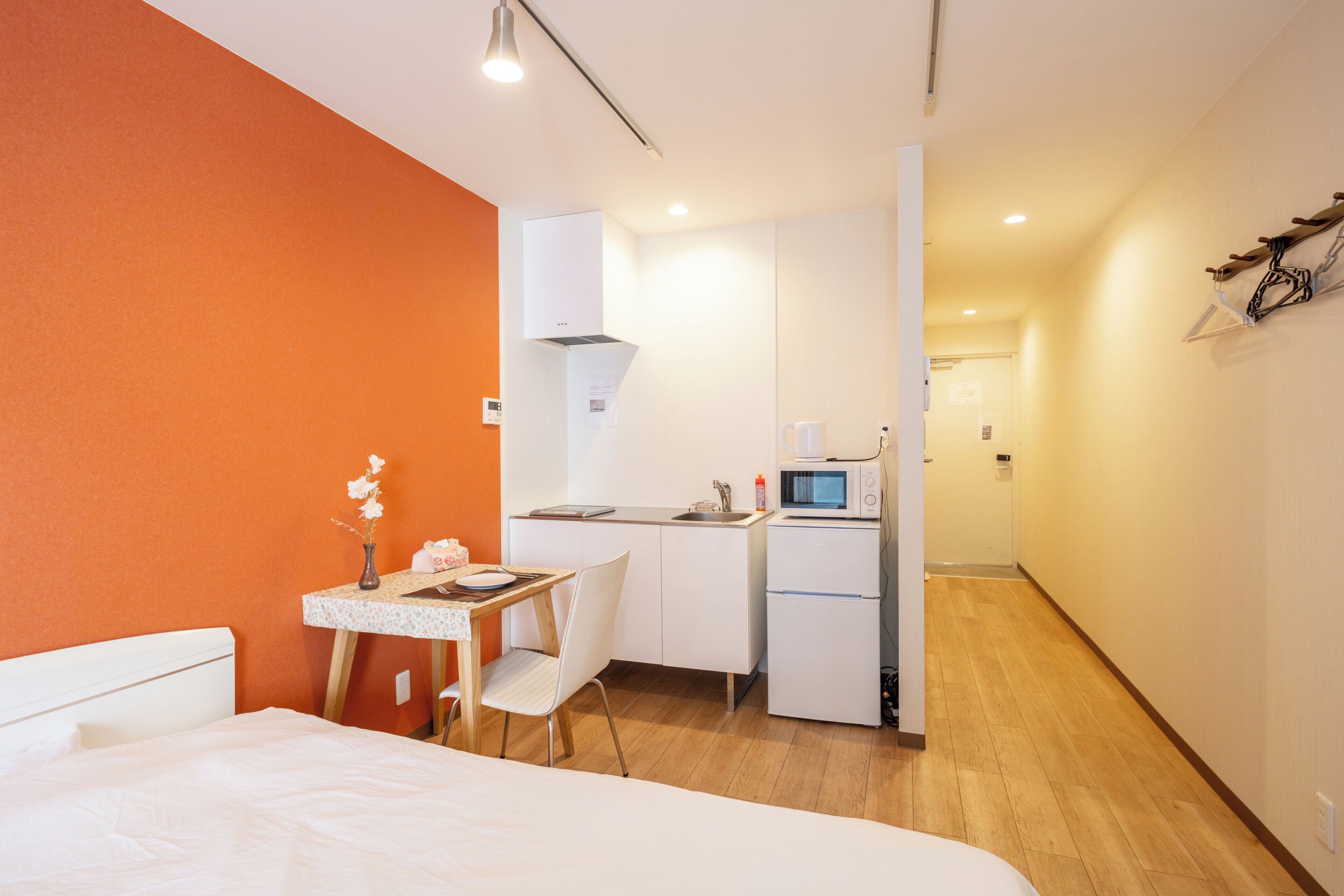 A hotel room with a bed, a small desk with a chair, a kitchenette, and a bathroom visible in the background.