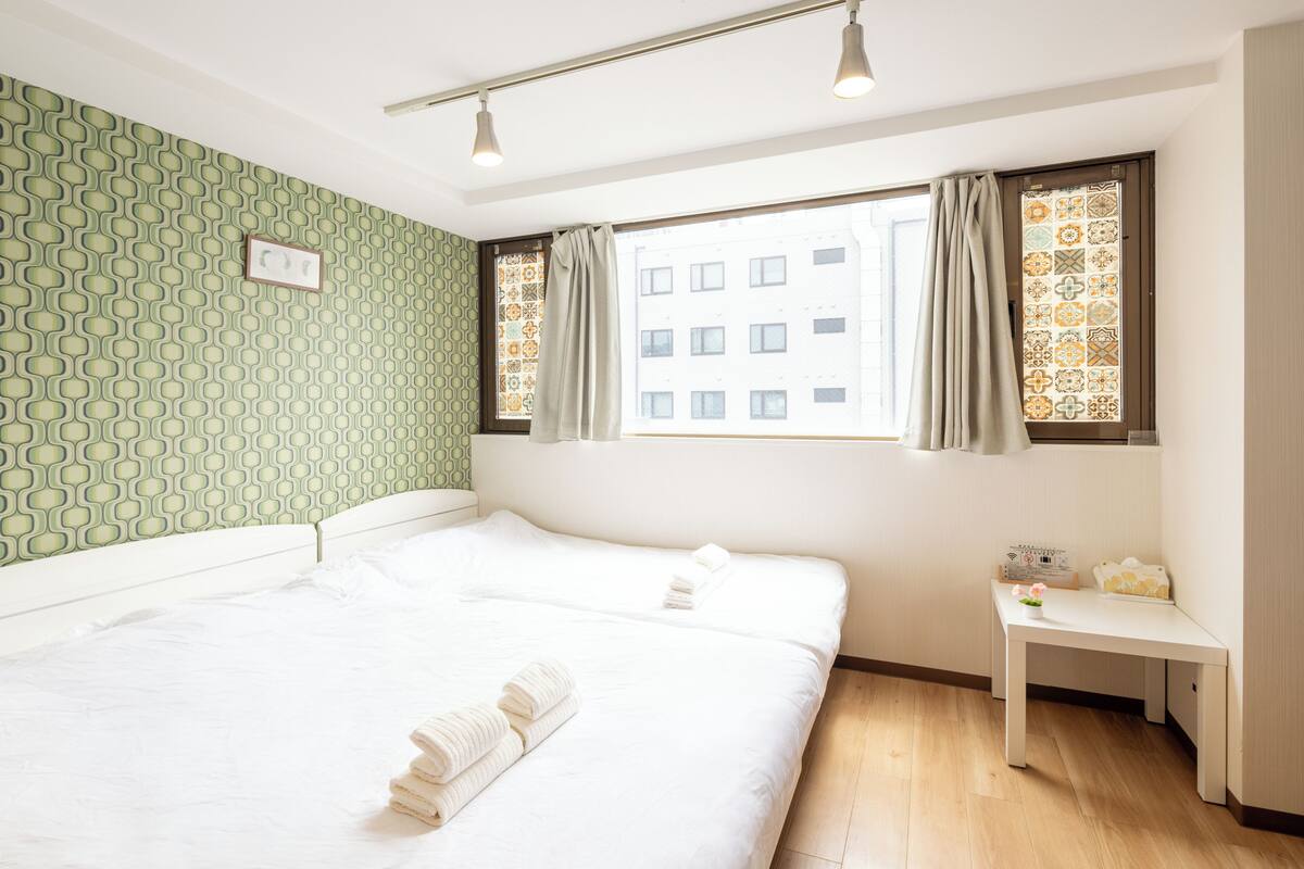 Quarto de hotel com duas camas, uma mesa pequena e uma janela com cortinas estampadas.