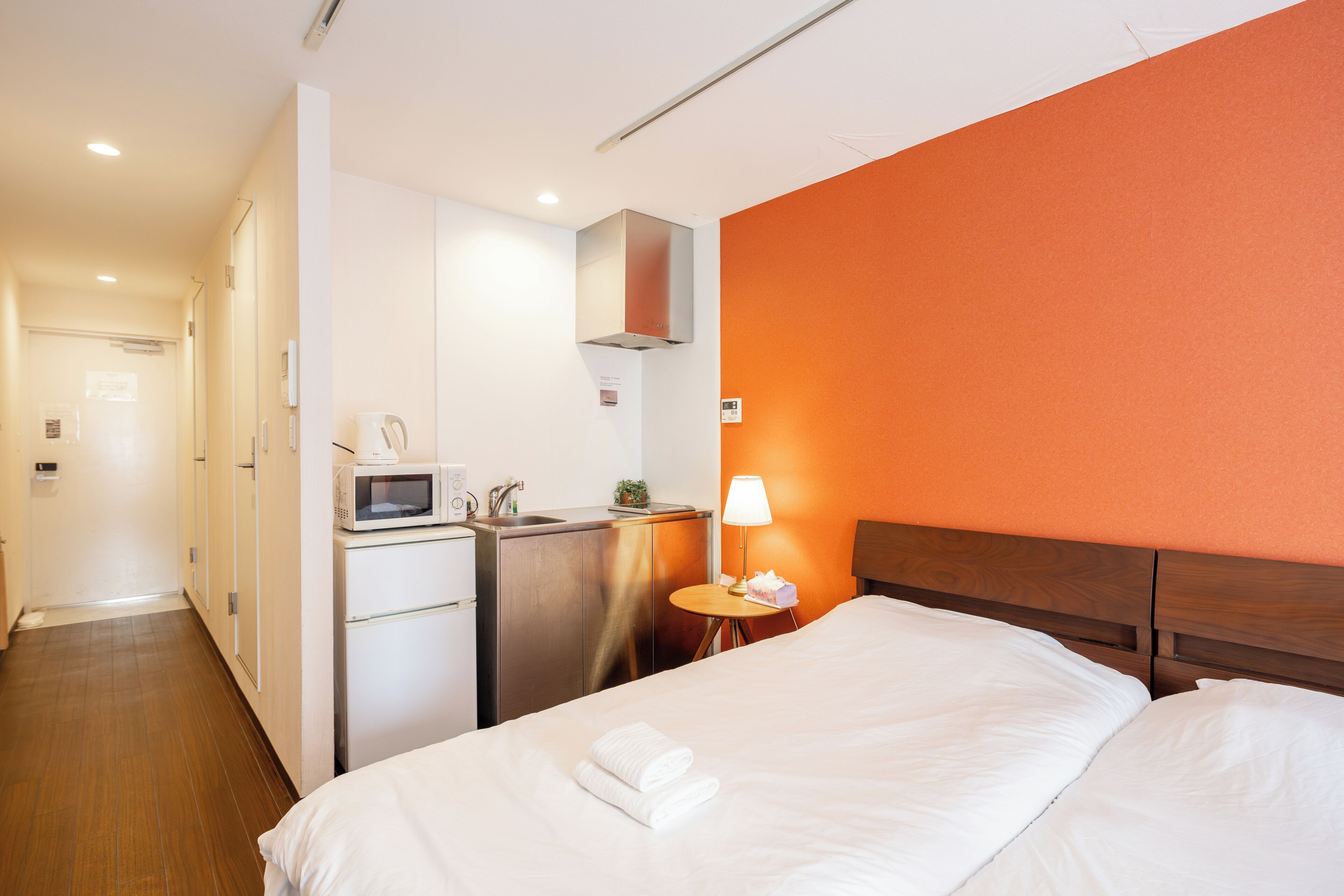 Hotellihuone, jossa on sänky, työpöytä, jossa on mikroaaltouuni, lamppu ja pieni pöytä, jonka päällä on paperinenäliöpakkaus.