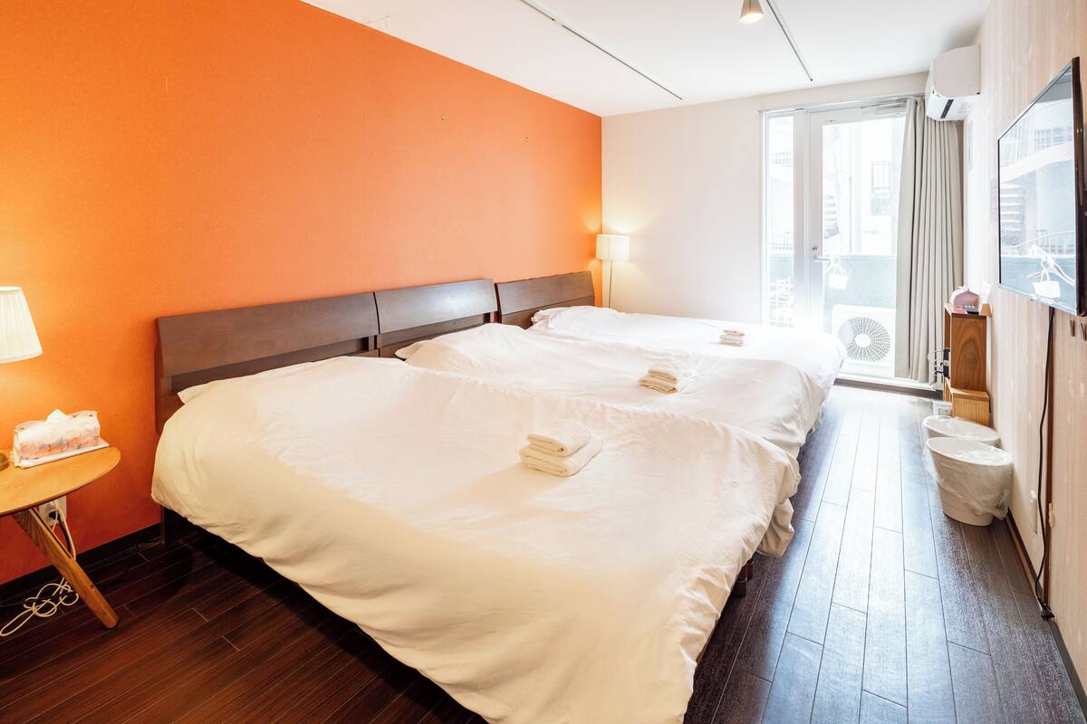Quarto de hotel com duas camas, uma mesinha com luminária, um ventilador e uma televisão fixada na parede.