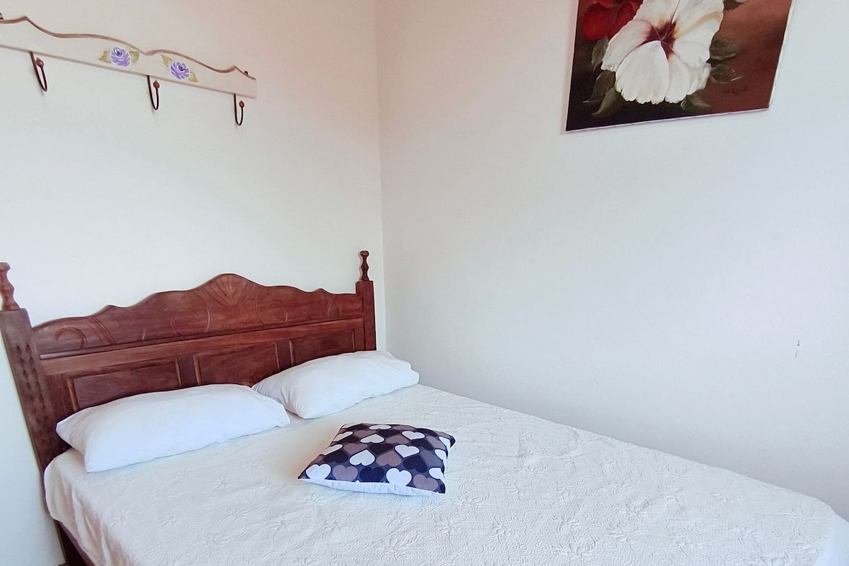 A neatly made bed with a patterned bedspread and two pillows, a wooden headboard, and a wall-mounted artwork of flowers.