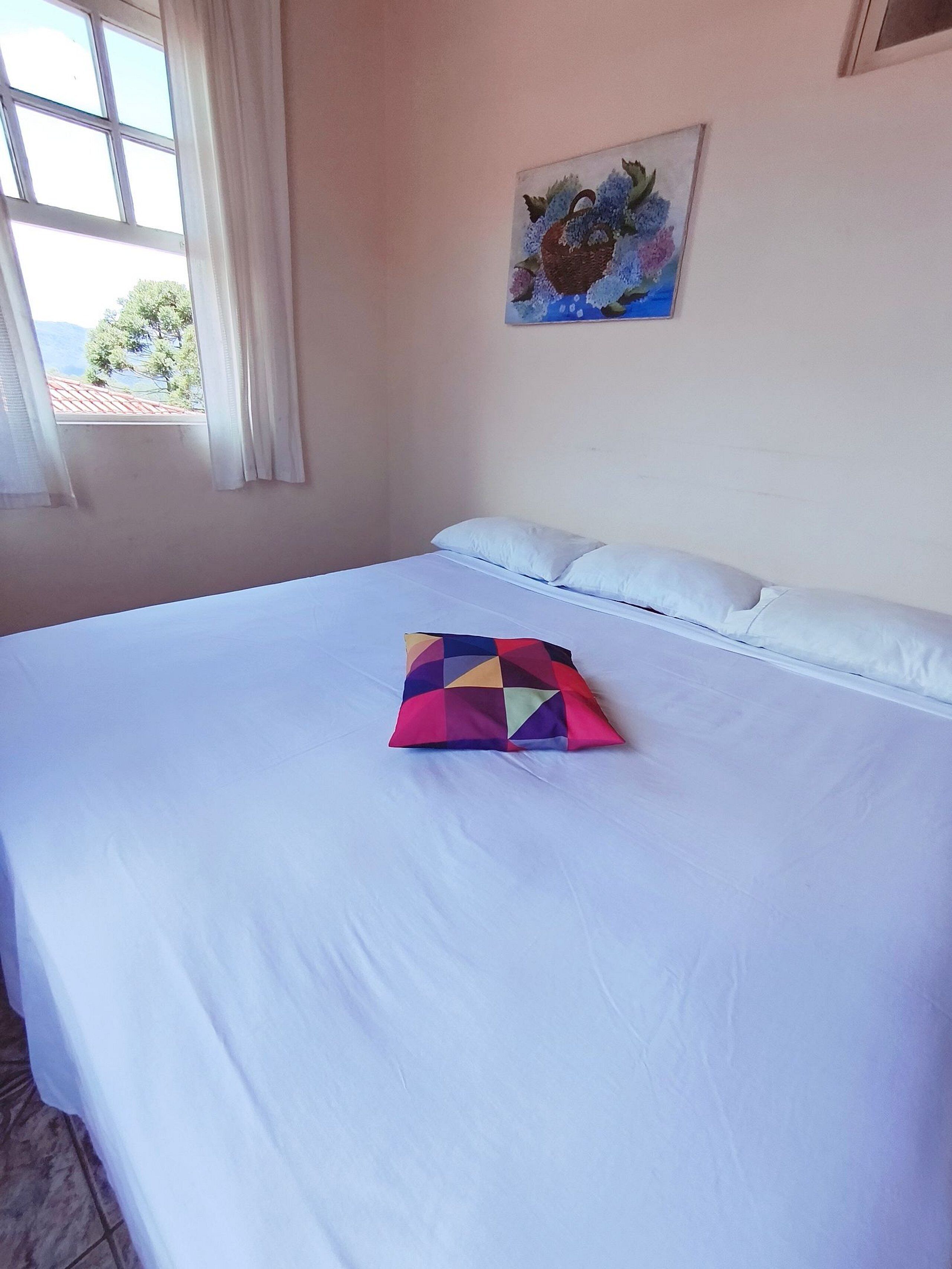 Un lit bien fait, avec du linge de lit blanc et un coussin aux motifs géométriques colorés, un tableau au mur et une fenêtre avec des rideaux.