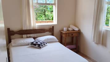 Een net opgemaakt bed met een houten hoofdbord, een klein nachtkastje met een lamp en een raam met gordijnen.