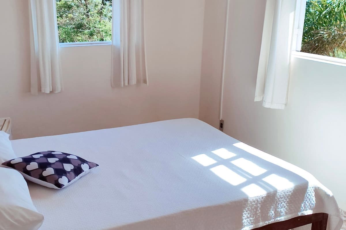 A single bed with a white mattress and a heart-patterned pillow, a wooden headboard, and a window with white curtains.