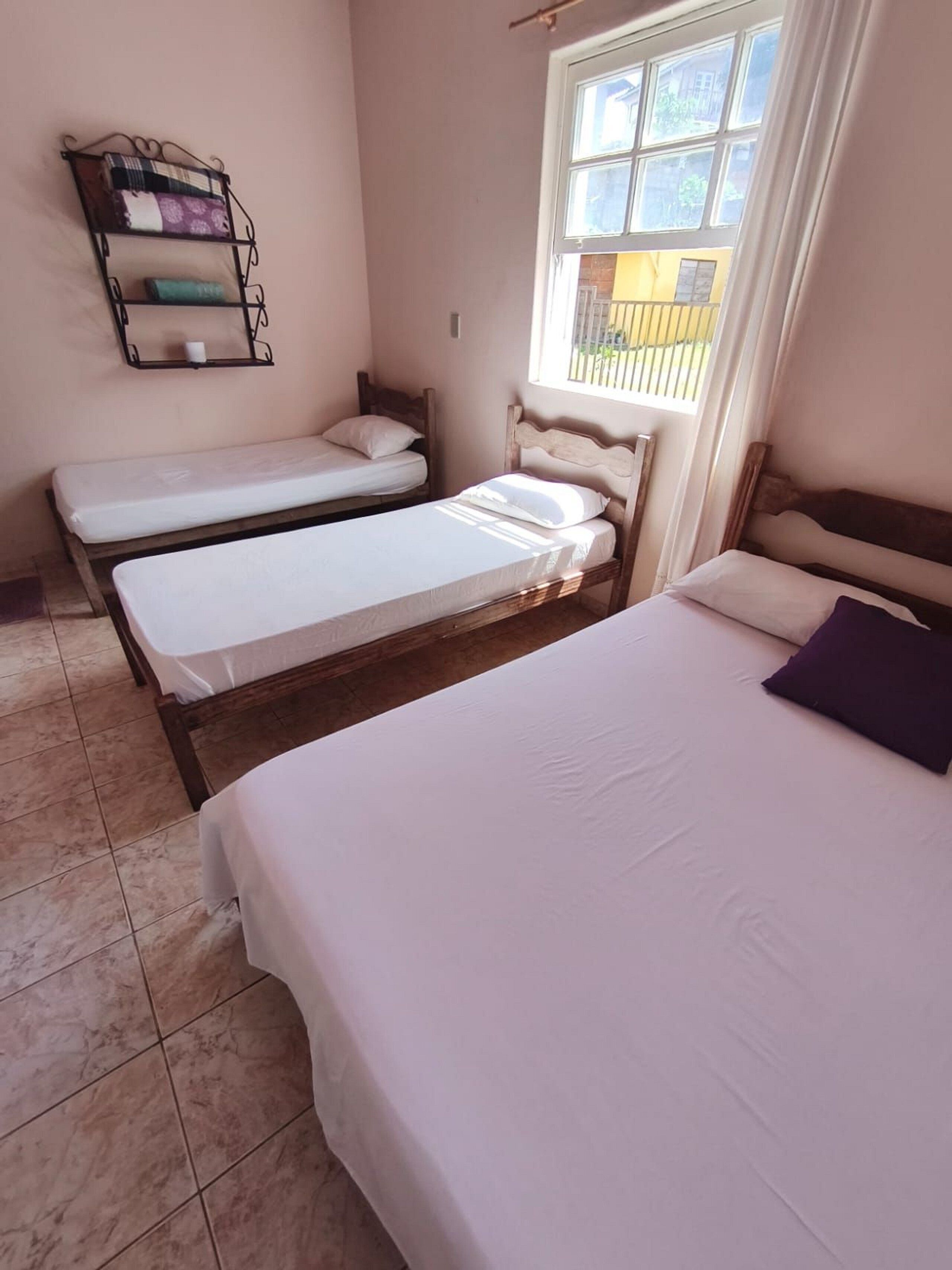 A room with two single beds, a window with blinds, a shelf with books, and a tiled floor.