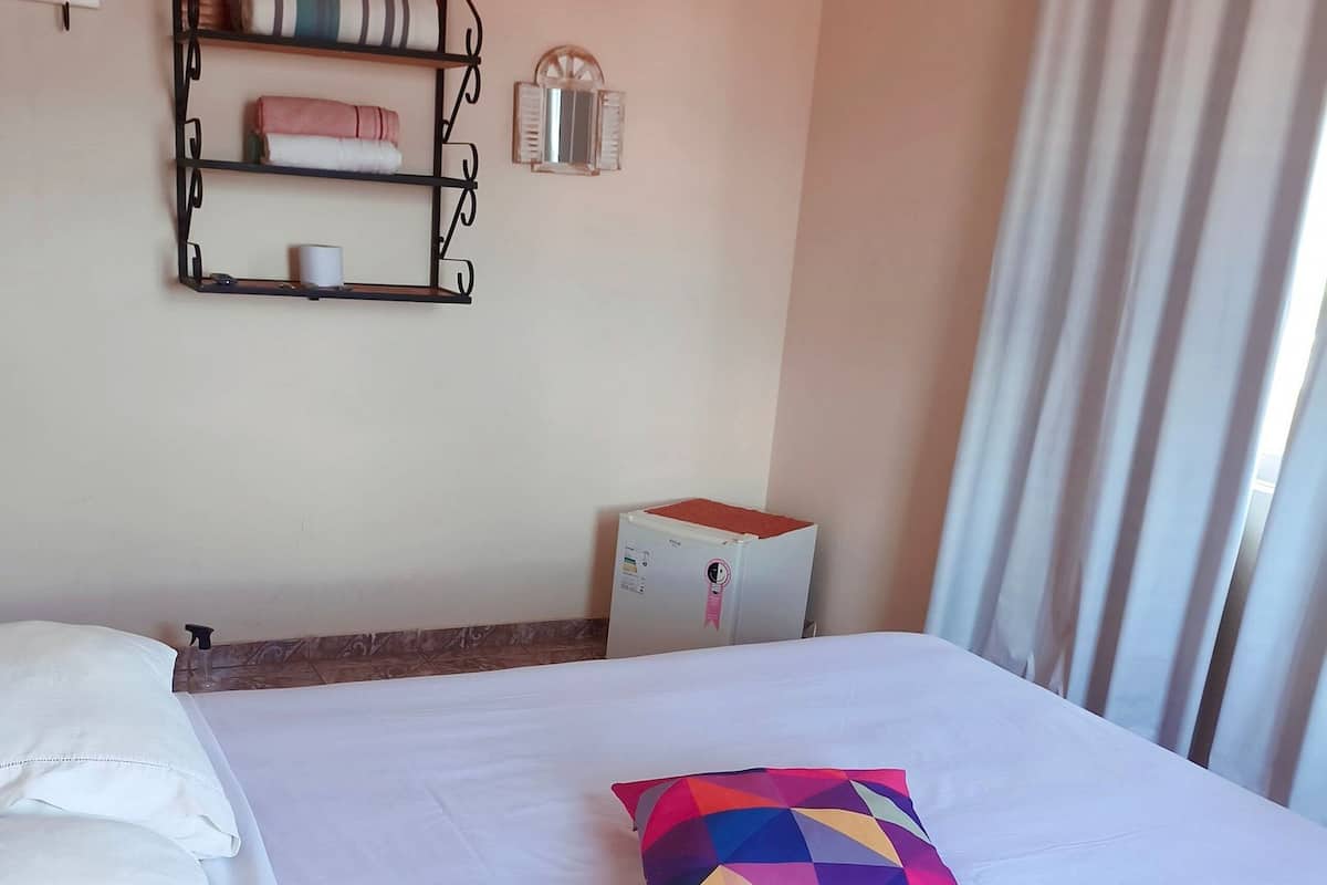 A neatly made bed with white linens, a colorful geometric pillow, a wall-mounted shelf with towels, a small refrigerator, and a window with curtains.