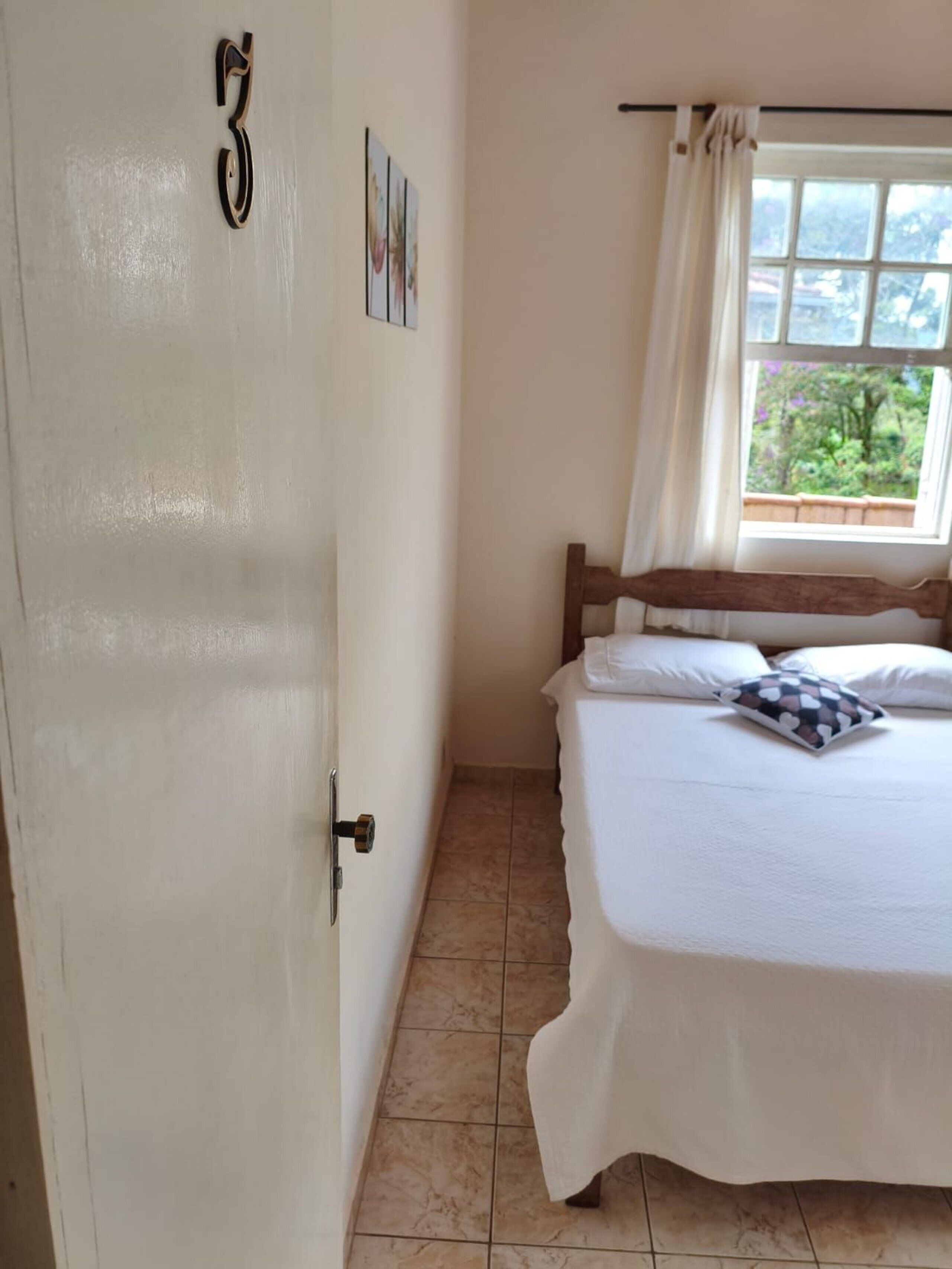 Quarto com cama, janela com cortinas, porta e quadro pendurado na parede.