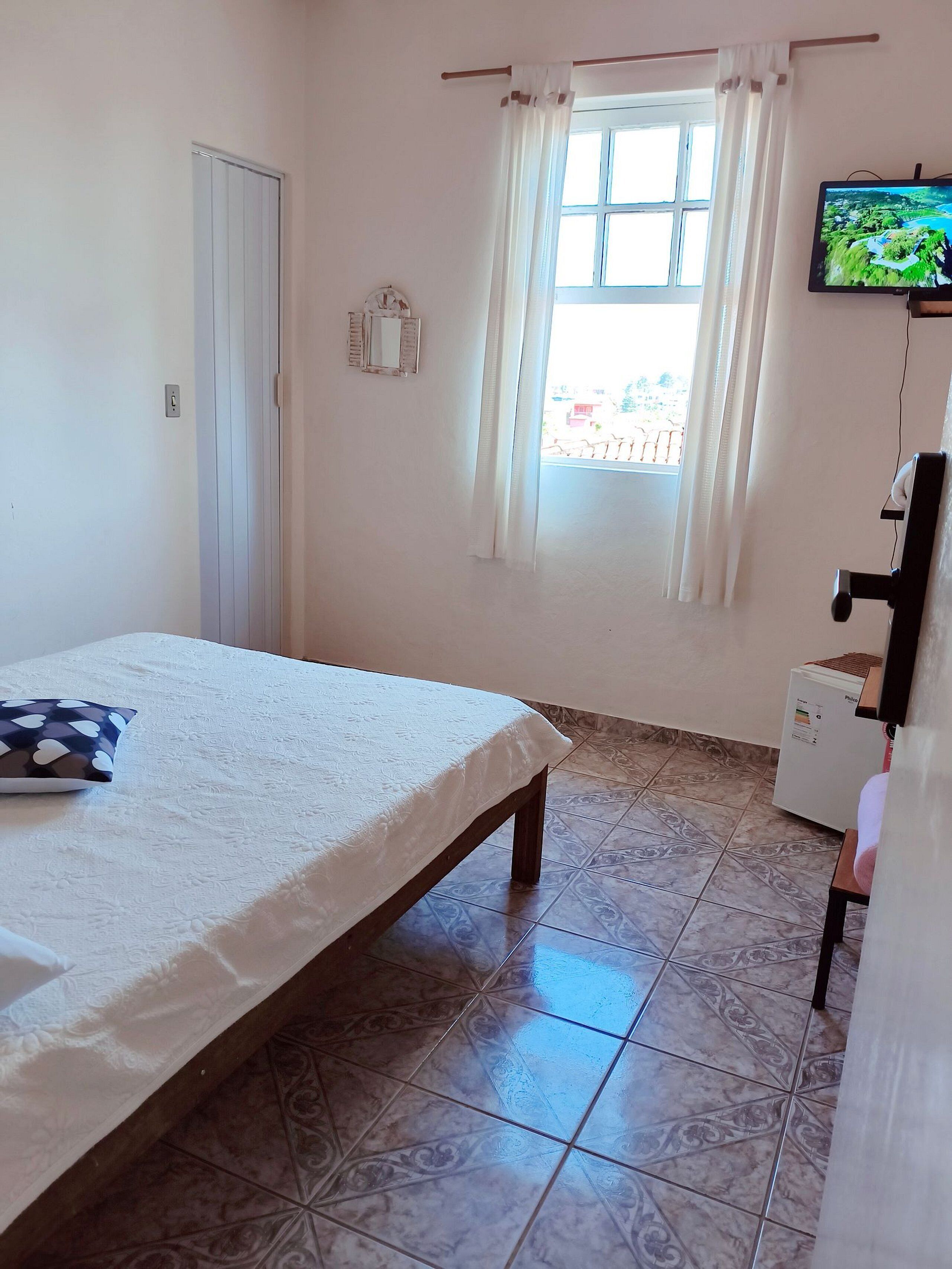 Ein Schlafzimmer mit einem Bett, einem an der Wand befestigten Fernseher, einem Fenster mit Vorhängen und einem gefliesten Boden.