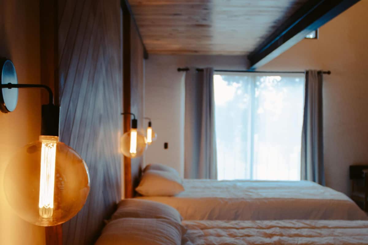 A room with two beds, wooden walls, a wooden ceiling, and a window with curtains.