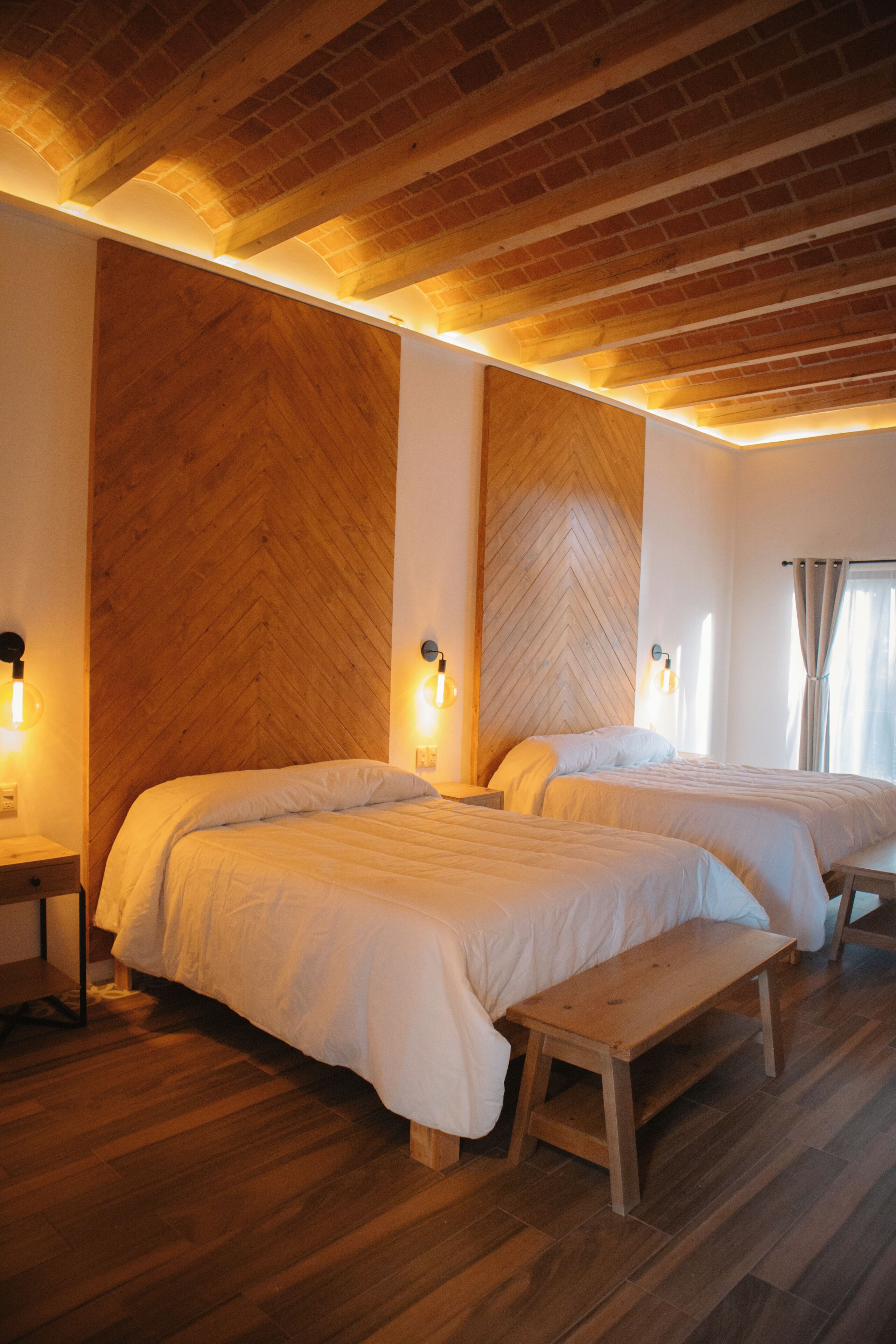 A room with two beds, wooden flooring, a bench, and a herringbone-patterned wall.