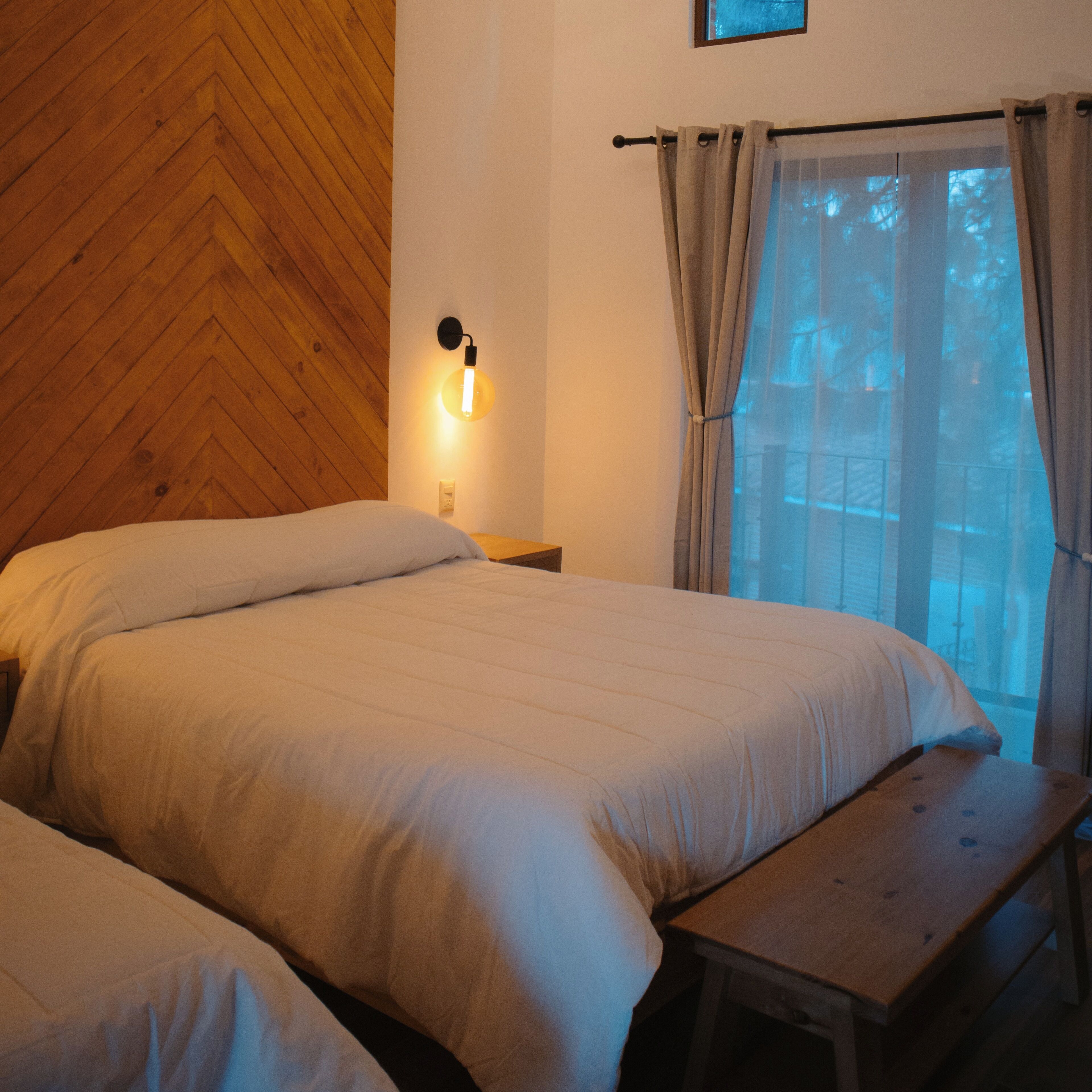 A bedroom with two beds, a nightstand, a bench, a wall-mounted light, and a window with curtains.