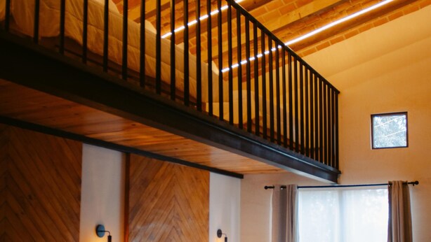 A bedroom with two beds, a wooden bench, a staircase, and a window with curtains.