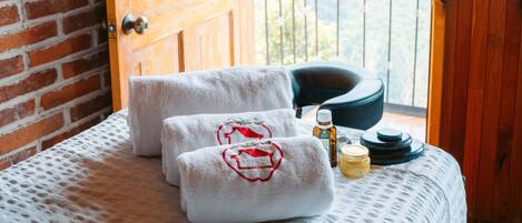 A room with a wooden door, a bed with a white textured bedspread, two towels with red embroidery, a bottle of olive oil, and a small bowl on the bed.