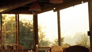 A dining area with wooden chairs and tables, large windows, and a view of trees outside.