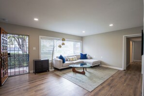 Un salon avec un canapé, une table basse en verre, un lampadaire et une vue sur l’extérieur, visible à travers une porte.