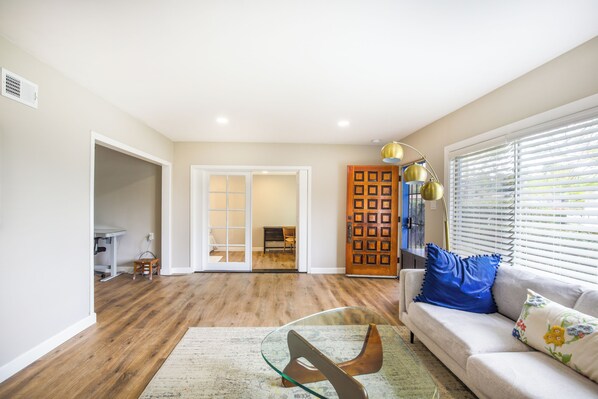 A living room with a sofa, a glass coffee table, a wooden floor, a door, and a window with blinds.