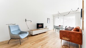 A modern living room with a light blue armchair, a wooden TV stand, a flat-screen TV, a red sofa, and a dining area with a table and chairs.