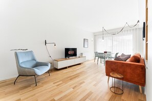 Ein modernes Wohnzimmer mit einem hellblauen Sessel, einer Holz-TV-Konsole, einem Flachbildfernseher, einem roten Sofa und einem Essbereich mit Tisch und Stühlen.