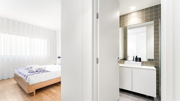 A modern hotel room with a wooden bed, a white bathroom with a large mirror, and a view of the city through the window.