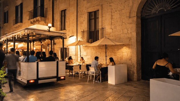 Un área de comedor al aire libre con mesas y sombrillas, un carrito blanco, y un edificio de piedra con ventanas y puertas de arco.