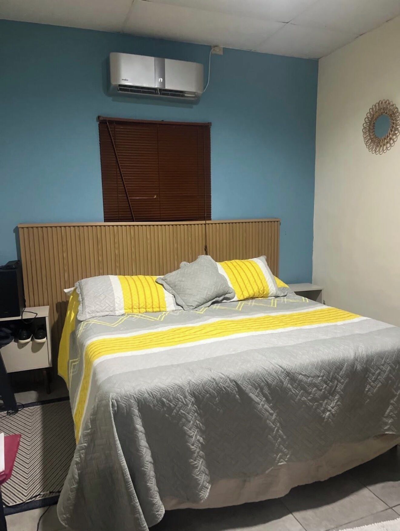 Une chambre à coucher comprenant un lit, une table de chevet, un bureau, un climatiseur, une fenêtre avec des stores et un élément décoratif mural.