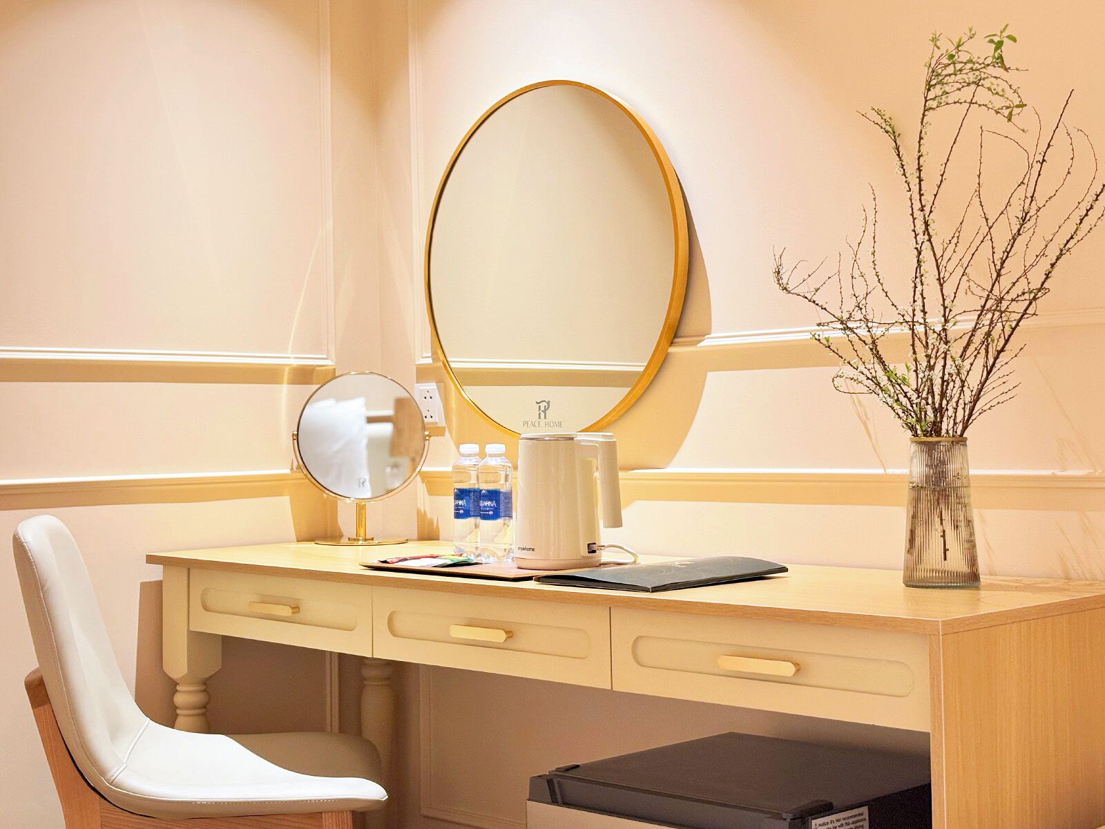 Habitación de hotel con un escritorio de madera, un espejo, un florero con ramas, una silla y un pequeño cubo de basura.