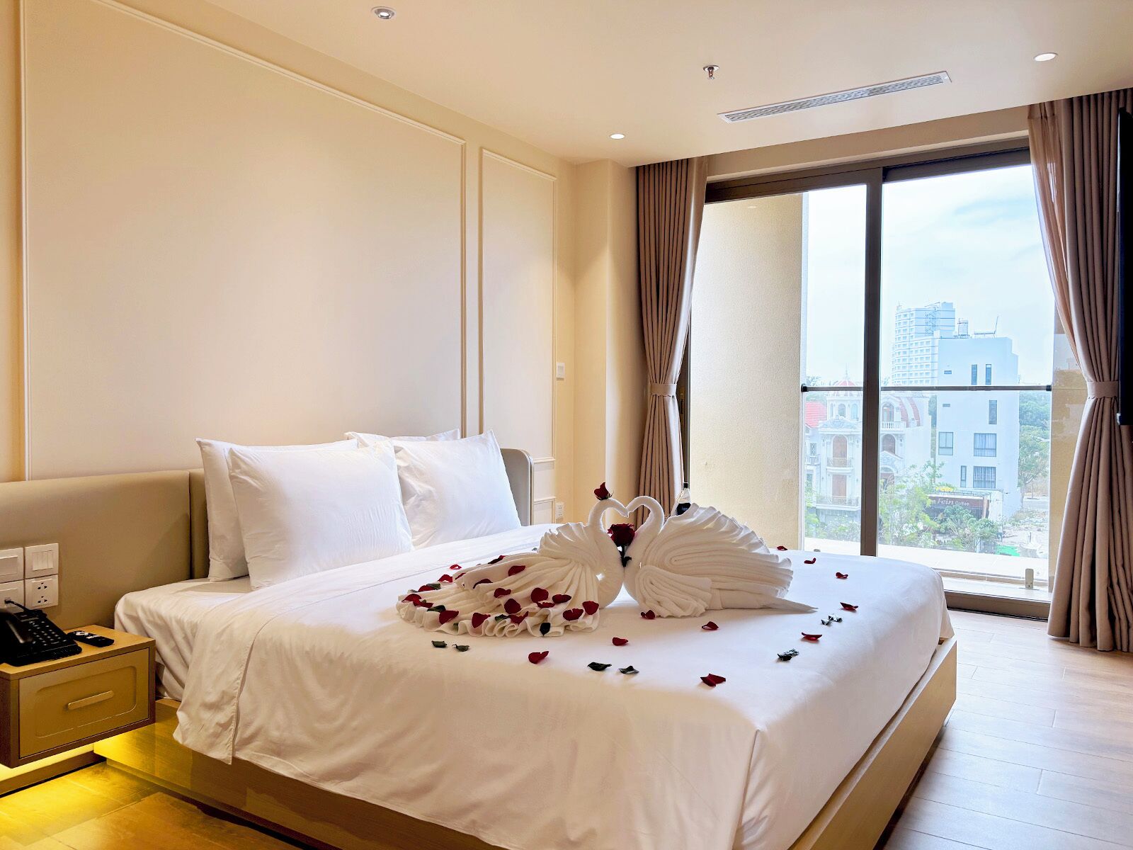 A hotel room with a large bed, a bedside telephone, and a view of buildings through the window.