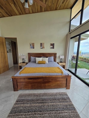 A spacious bedroom with a large bed, wooden headboard, and a view of the outdoors through large windows.