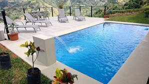 Et rektangulært swimmingpool med klar, blå vandoverflade, omgivet af en flisebelagt terrasse. Der er liggestole og et bord på terrassen, og plantekasser er placeret rundt i poolområdet.