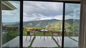 Et værelse med en skydedør, der fører ud til en udendørs terrasse med et bord og stole, med udsigt over et bjergrigt landskab.