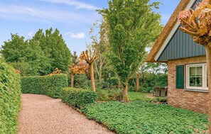 Een grindpad leidt naar een bakstenen huis met een groen raam, omringd door verzorgde hagen en bomen.