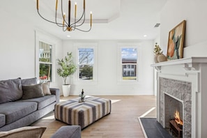 Ein modernes Wohnzimmer mit Kamin, grauem Sofa, Pouf mit Muster und einem großen Fenster.