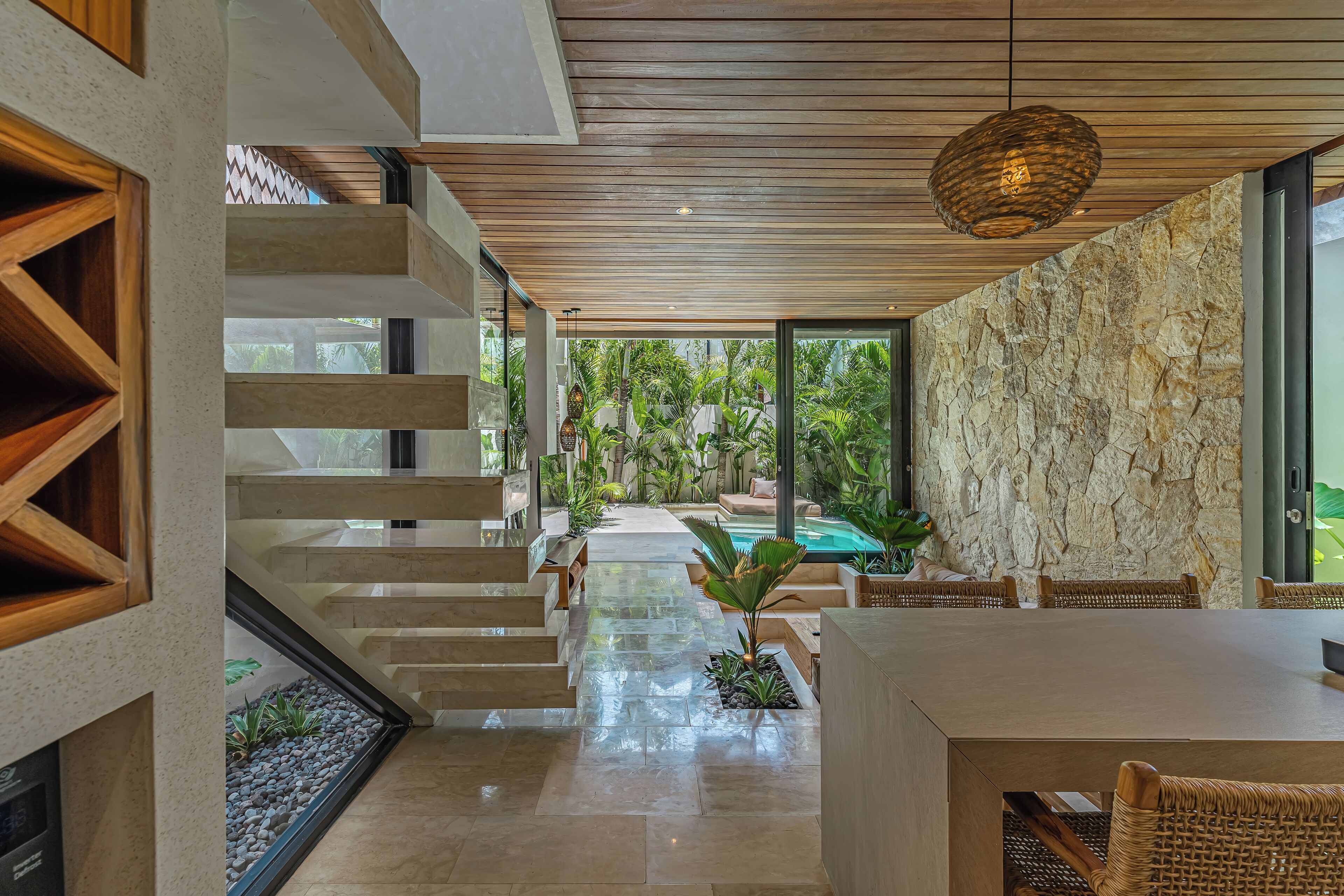 Un interior moderno con una escalera que lleva a una zona de piscina, paredes de piedra y un comedor con muebles de mimbre.
