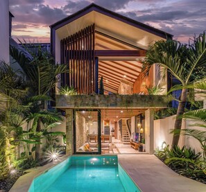 Una casa moderna de dos pisos con piscina, terraza de madera y plantas tropicales.