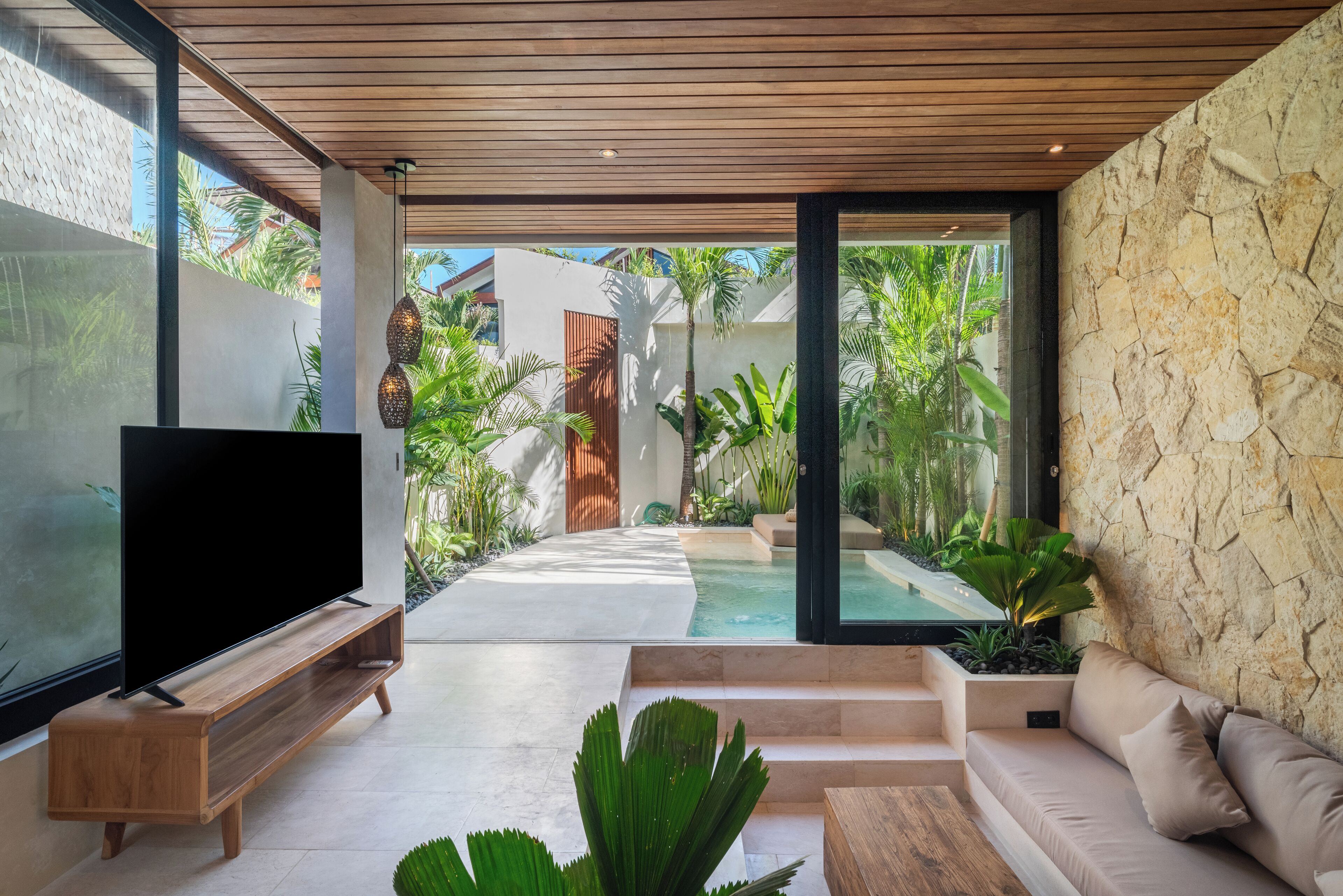 Una sala de estar moderna con un televisor de pantalla plana sobre un soporte de madera, un sofá color beige y una pared de piedra.