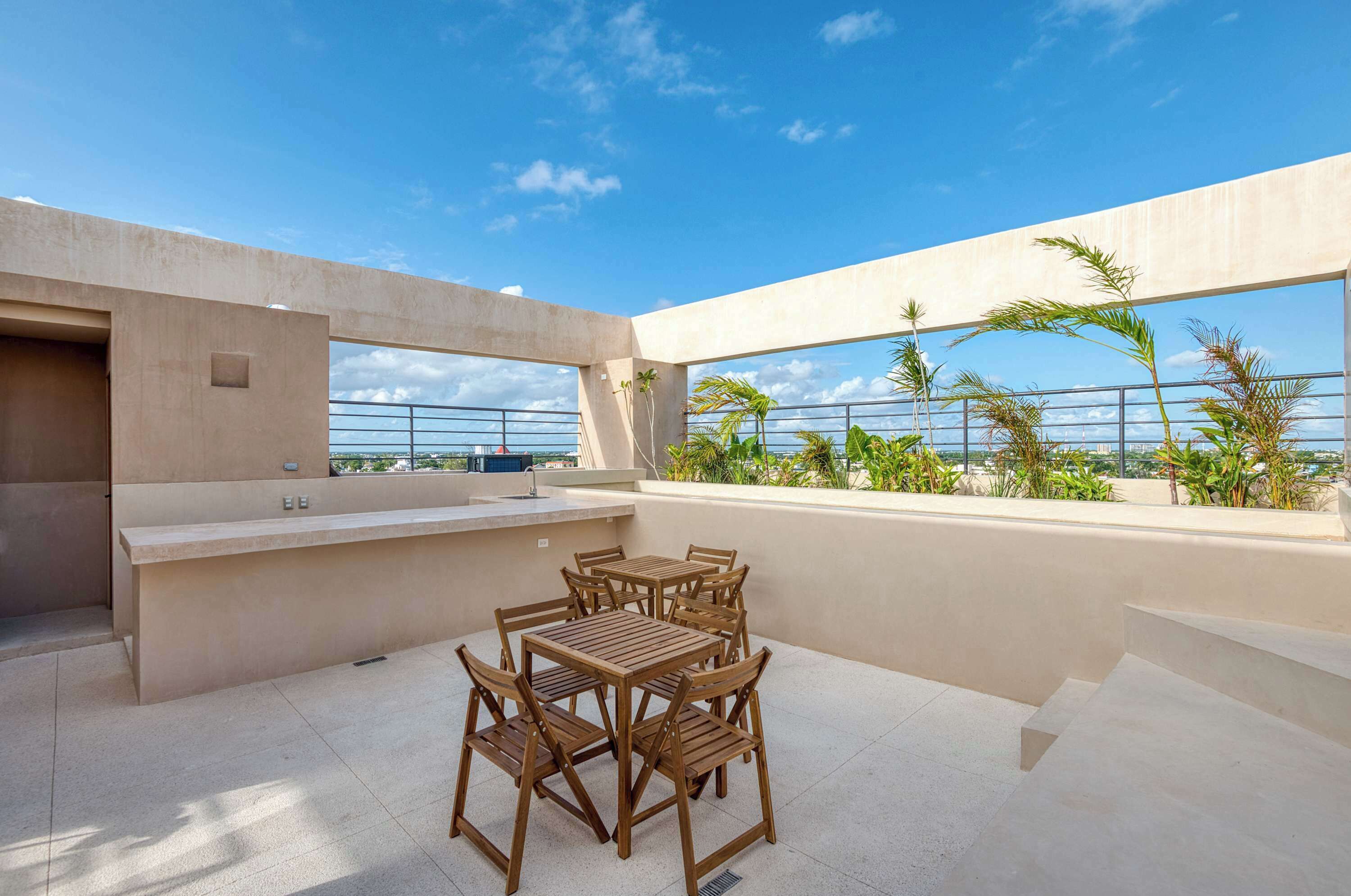 A rooftop terrace with a wooden table and chairs, a small kitchen area, and a view of palm trees and the ocean.